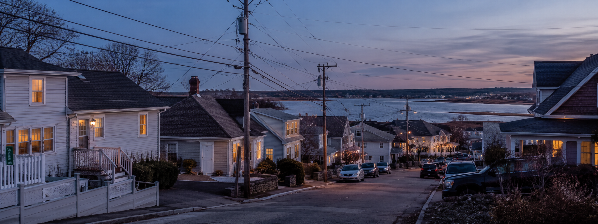 A wide horizontal shot of a Rhode Island shoreline neighborhood at peak utility load hour: late afternoon to early evening, lights coming on inside houses, distribution poles and a pole-mounted transformer in frame, hint of Narragansett Bay in the distance